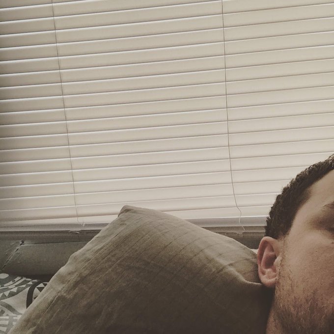 A man's face is partially visible on the right side of the frame, resting against a brown pillow with white window blinds in the background.