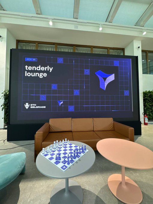 A lounge area at ETH Belgrade features a large screen displaying 'tenderly lounge' with an ETH Belgrade logo, accompanied by a chess board set up on a round table next to a tan sofa.