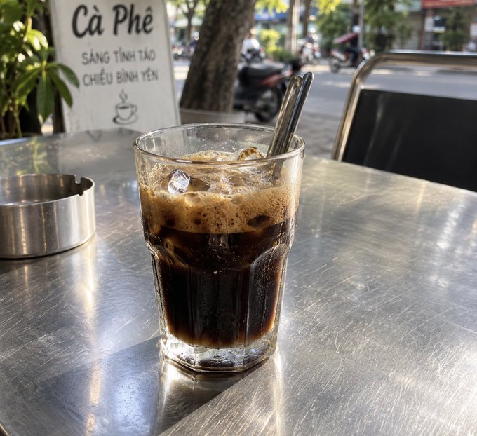A glass of Vietnamese iced coffee (cà phê) with ice and a metal spoon on a stainless steel table at an outdoor cafe, with a sign reading 'Cà Phê SÁNG TỈNH TÁO CHIỀU BÌNH YÊN' and a street with motorcycles in the background.