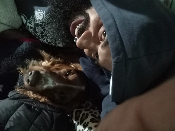 A dimly lit selfie of a man with a beard lying next to a small brown and white dog.