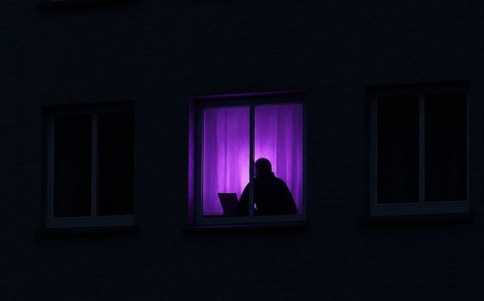 A dark nighttime photo of a building's facade with three windows; the central window is illuminated with purple light, revealing the silhouette of a hooded figure using a laptop.