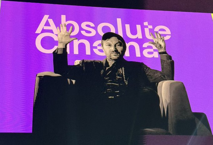 I cannot identify the individual in the image. However, I can describe what I see: a person wearing a dark outfit and cap sits in a chair with both hands raised in the air, set against a purple backdrop displaying the text "Absolute Cinema" in large white letters.