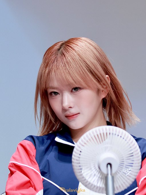A young woman with short blonde hair wearing a blue and pink jacket, looking toward the camera with a small fan in front of her; the watermark reads 'Kalavi8ka'.