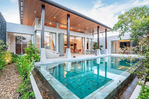 A luxurious modern villa features an infinity pool, glass walls, and a covered patio with a lounge area, surrounded by tropical landscaping.