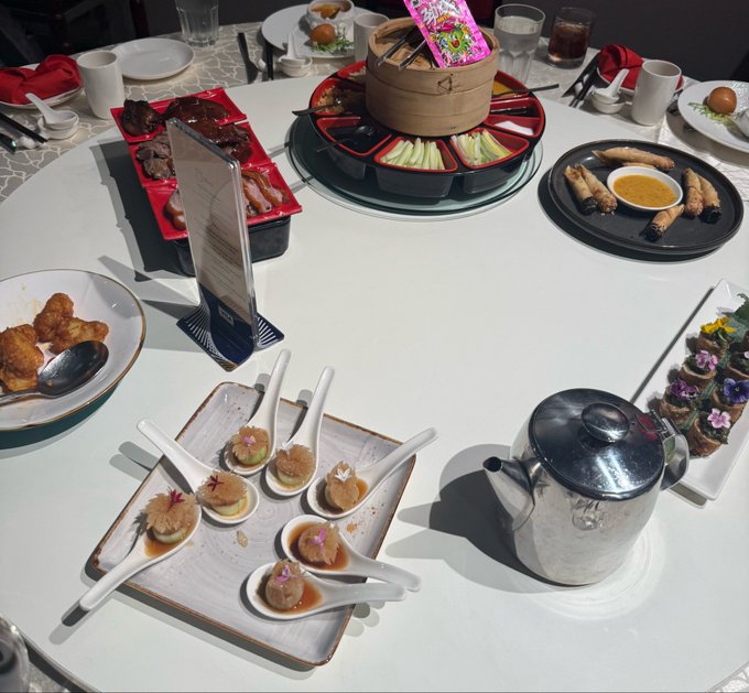 A round dining table set with a Chinese feast, including bamboo steamers of dim sum, spring rolls with dipping sauce, assorted meats on red plates, and spoons of appetizers with a silver teapot.