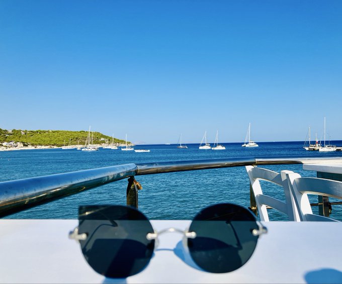 A first-person view from a boat looking out over blue ocean water with sailboats and a green coastline in the distance, with round sunglasses resting on the boat railing in the foreground.