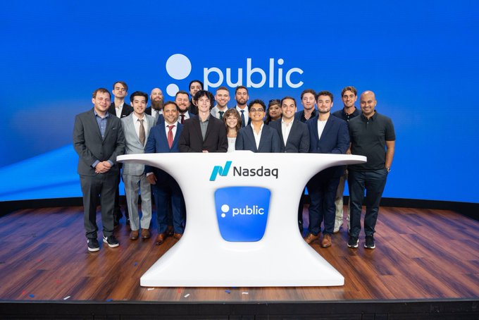 Group photo of approximately 15 people in business attire standing behind a white podium displaying the public logo and Nasdaq branding, with 'public' logo displayed on a blue backdrop, suggesting an IPO or Nasdaq listing event.