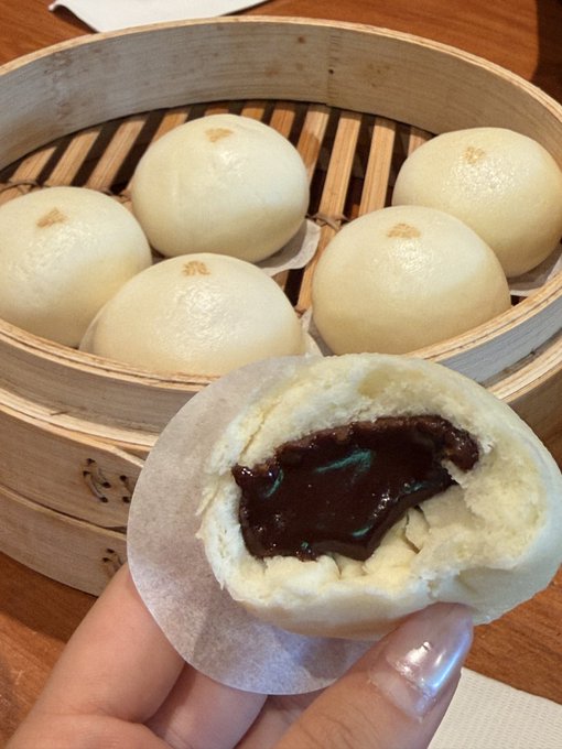 A hand holds a steamed bun (baozi) filled with red bean paste, with a bamboo steamer of additional buns in the background.