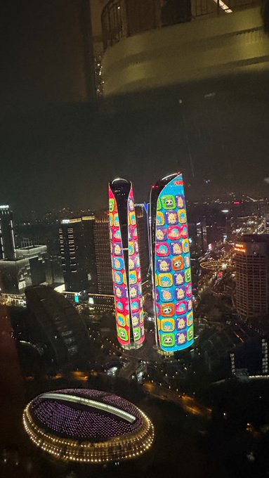 A nighttime cityscape view featuring two tall skyscrapers illuminated with colorful LED displays showing cartoon characters, set against an urban skyline.
