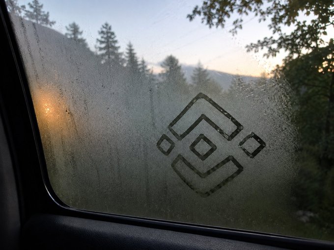 A foggy, condensation-covered car window with the Binance logo etched in the mist overlooks a mountain landscape at sunrise, evoking an "early morning" scene that aligns with the post's groggy wake-up call.