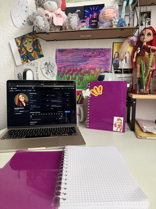 A desk setup featuring a laptop displaying a dark-themed interface, a purple notebook, and various colorful artwork, plush toys, and stationery on shelves in the background.