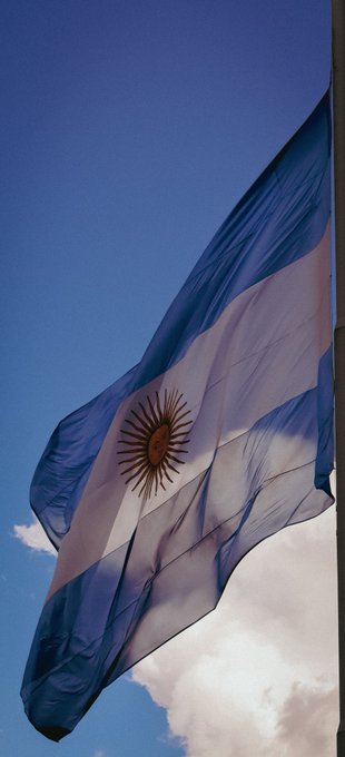 The flag of Argentina waving against a bright blue sky with scattered clouds, featuring its distinctive light blue and white horizontal stripes with the Sun of May in the center.