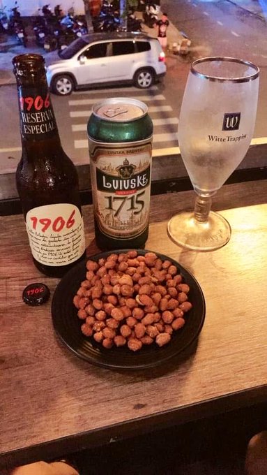 A bar setup on a wooden ledge overlooks a street with parked cars, featuring a bottle of '1906 Reserva Especial' beer, a can of 'Lüiske 1715' beer, a 'Witte Trappiste' chalice, and a small black plate of roasted nuts.