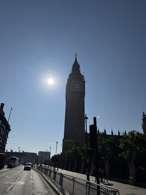 A sunlit street view of London's Elizabeth Tower (Big Ben) in silhouette against a bright blue sky, with the sun visible to the left of the clock tower.
