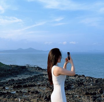 The image features a woman standing near the ocean, holding her cell phone up to take a picture of the beautiful scenery around her. The photo captures the vast expanse of water and the rocky shoreline where she stands. The caption "休假休假啊 可以慢慢定投btc了" suggests that this moment is a special one, possibly during an event or gathering involving Bitcoin transactions.