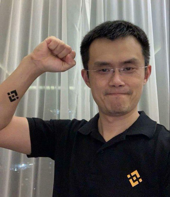 Changpeng Zhao (CZ), the former CEO of Binance, raising his fist to show a temporary Binance tattoo on his forearm, wearing a black polo shirt with the Binance logo.