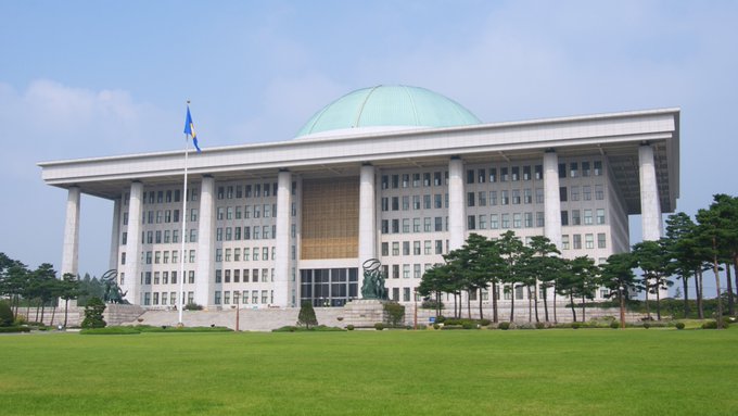 A large modern government building with a prominent green dome and a flag flying out front, set behind a well-manicured lawn.