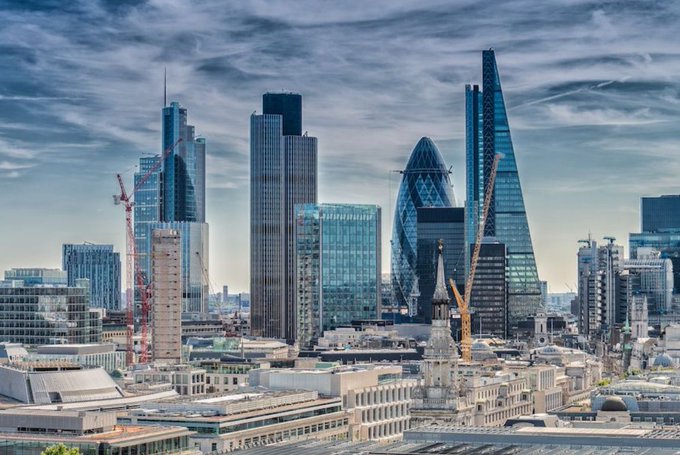 A daytime photograph captures a sprawling city skyline, prominently featuring London's financial district with its modern skyscrapers alongside older architectural styles under a cloudy sky.