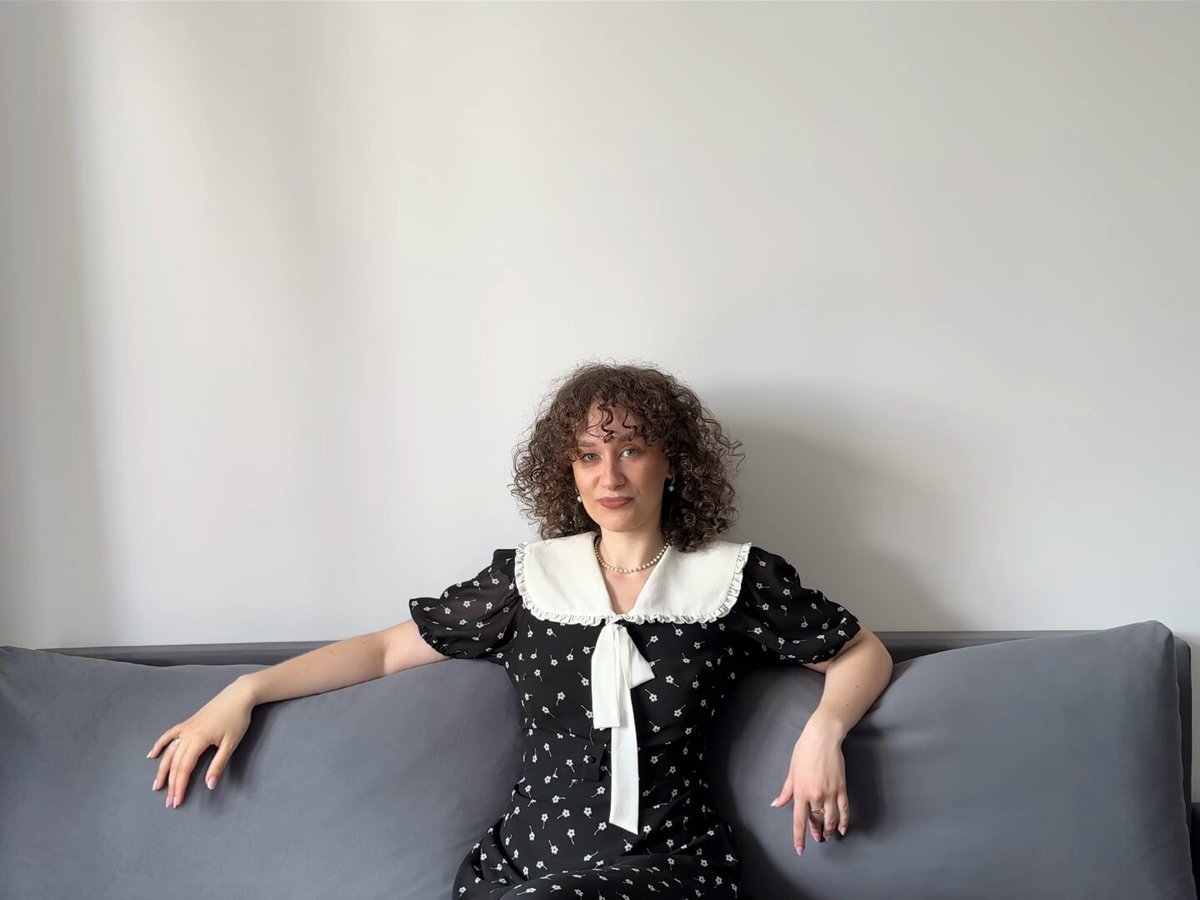 The image features a woman sitting on a couch with her legs crossed. She is wearing a black dress with white polka dots and has curly hair. The background of the image shows a wall, which could be part of a living room or bedroom setting. There are no texts present in the image, so it's unclear if there is any information about the post related to this image.