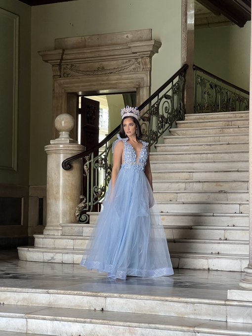 A woman wearing a tiara and an elegant light blue gown stands at the base of a grand marble staircase.