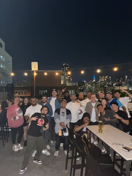 A group photo of around 20 people posing together on a rooftop bar at night with city skyscrapers and string lights in the background.