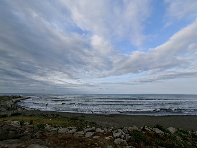 A photograph of a calm beach at dusk or dawn, with rocky shoreline, gentle waves, and a cloudy sky.