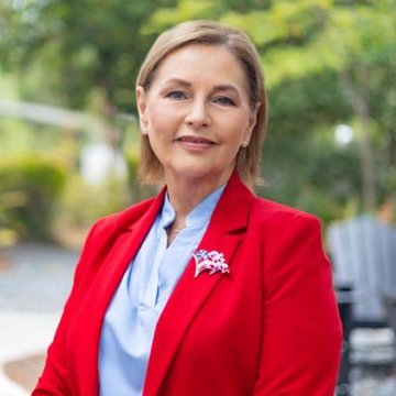 A confident woman with short blonde hair, wearing a red blazer over a light blue blouse and a sparkling butterfly brooch, stands outdoors with green foliage in the background. Her poised, professional appearance aligns with the post's caption of a U.S. Senator speaking about supporting market structure legislation.