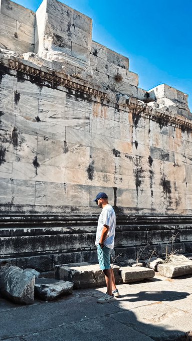 A man wearing a white t-shirt, green shorts, and a cap standing in front of a massive ancient marble wall under a clear blue sky.