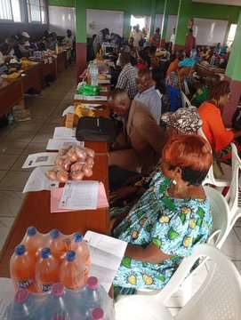The image shows a group of people sitting at tables with papers and bottles, possibly discussing or selling items. The scene appears to be set up for an event like the Traditional Monthly Periodic Market Day held by the community. There are several people present in the room, including some who seem to be actively engaged in conversation while others appear to be focused on their tasks. 

The tables have various items such as bottles and papers, which could be related to the market day activities or other purposes. The image also contains a caption that reads "Focus on what is in the image and how it relates to the post". This suggests that the image might be used for promotional or educational purposes, highlighting the importance of community events like the Traditional Monthly Periodic Market Day.
