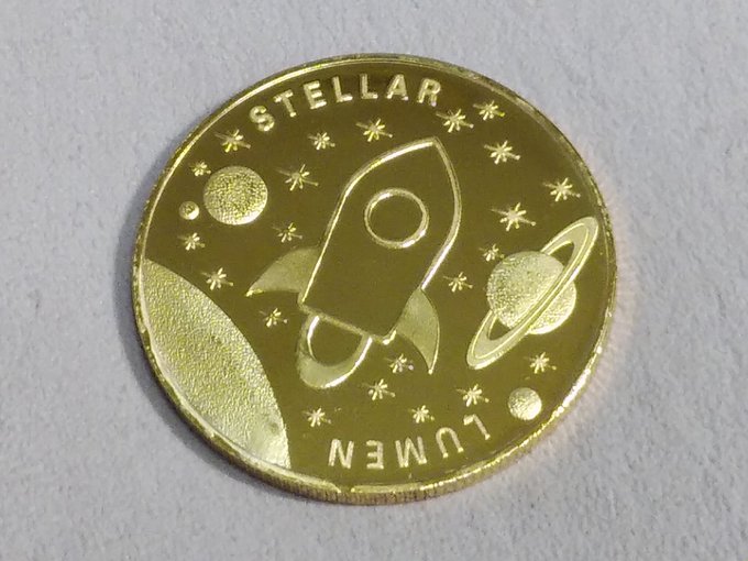 A commemorative gold-colored Stellar Lumens (XLM) physical coin featuring a rocket, planets, and stars with the text "STELLAR LUMEN," representing the cryptocurrency delivery mentioned in the post.