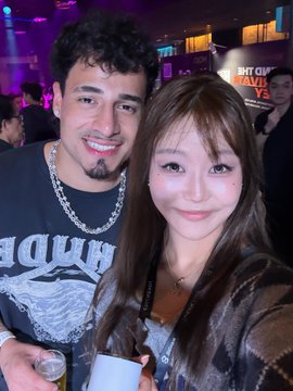 A selfie-style photo of a smiling young man with dark curly hair wearing a chain necklace and a young woman with long dark hair, posing together in a crowded indoor venue with purple lighting.