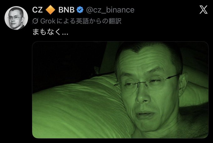 A night-vision photo shows CZ (Changpeng Zhao), founder of Binance, lying in bed wearing glasses with a tired expression, accompanied by a tweet from his @cz_binance account with the Japanese text "まもなく…" (meaning "soon…"). The image supports the breaking news headline by suggesting CZ is hinting at an imminent significant development in the cryptocurrency space.