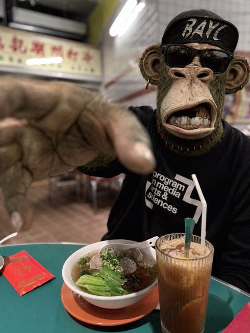 A person wearing a realistic monkey mask, sunglasses, and a BAYC (Bored Ape Yacht Club) cap, pointing at the camera while seated at a table with a bowl of noodle soup and a glass of iced tea.