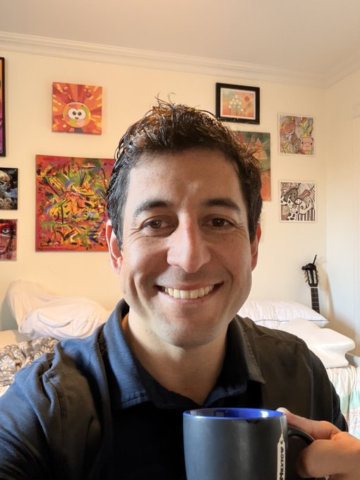 A smiling man holding a dark blue mug, sitting in a room with various colorful paintings and artwork on the wall behind him.