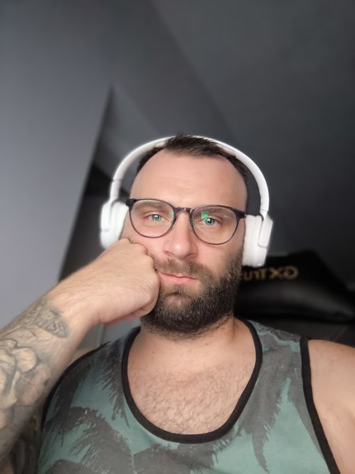 The image features a man wearing glasses, headphones, and holding up his hand over his face with one of them touching his mouth. The background includes a gray wall and the text "day 700 of posting selfie until $ZK hits 0.011" which appears to be part of a series of posts or pictures showcasing a similar setup in various locations around the world.