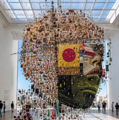 A large suspended mixed-media sculpture of a face made from numerous small components displayed in a bright museum atrium with people viewing it below.