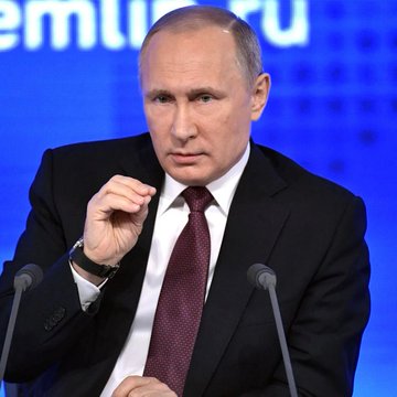 A man in a dark suit and maroon tie gestures while speaking at a podium with microphones, set against a blue backdrop displaying partial text including "erm" and "Ru." The image relates to the post as the new crypto registration rules in Russia are part of a broader regulatory framework being introduced under his administration.