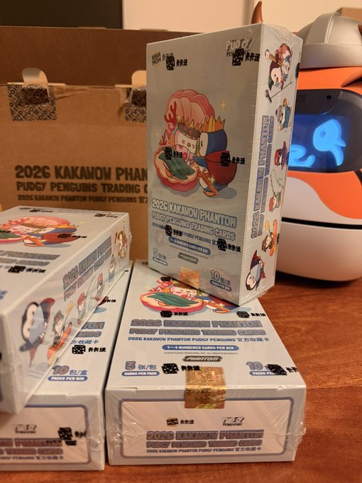Several sealed boxes of '2026 Kakamon Phantom Pudgy Penguins Trading Cards' stacked on a table, with a Pudgy Penguins branded helmet visible in the background.