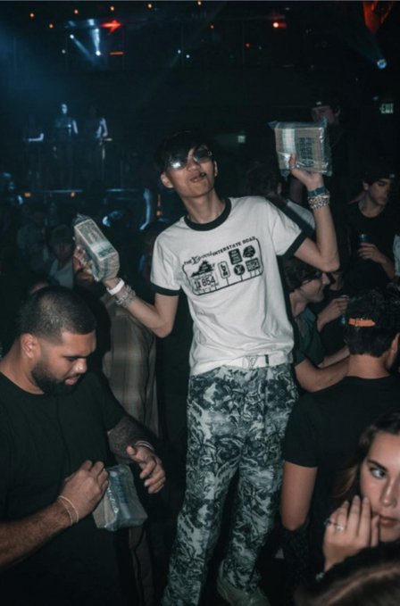 A man wearing sunglasses, a ringer t-shirt, and floral pants stands in a crowded club, holding up stacks of cash in both hands.