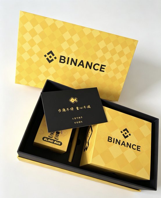 The image features a small box with the word "binance" written on it, along with some other text. The box appears to be made of cardboard or paperboard material and has a yellow background. It contains two small items - one is a pen, while the other is a card. The pen can be seen lying next to the card in the box.