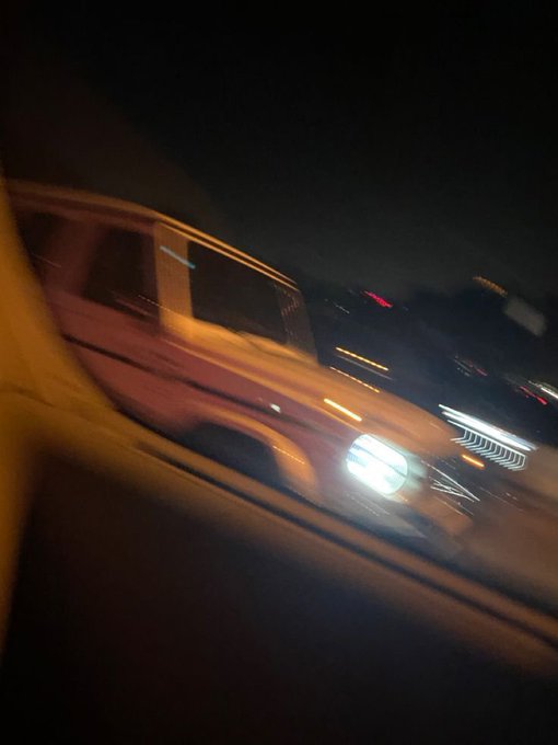 A blurry nighttime photograph taken from inside a vehicle, showing a white SUV or truck passing by with streaking lights from motion blur.