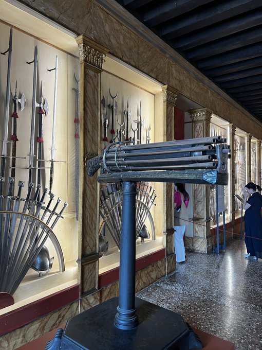 Interior of a museum armory displaying historical weapons like swords, spears, and a multi-barreled firearm mounted on a pedestal, with visitors observing the exhibits.