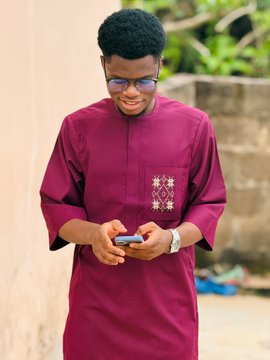 A young man wearing glasses and a maroon traditional embroidered tunic smiles while looking down at a smartphone.