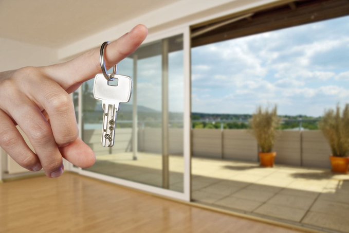 A hand dangles a set of keys in the foreground, with a bright, spacious empty home featuring a sunlit balcony and distant landscape visible behind. The image directly illustrates the post's core advice—encouraging investors to take profits during bull market cycles and convert crypto gains into real, tangible assets like property.
