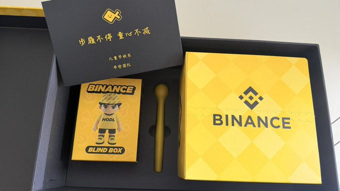 The image features a small toy binance trading card set, which includes a trading card game called Binance Blind Box. The cards are yellow with black text and illustrations of a boy holding a pen and a book titled "Binance Blind Box". The box is placed on top of another box that has the word "binance" written in Chinese characters. The post also mentions that there's a small toy binance trading card set, which includes a trading card game called Binance Blind Box.