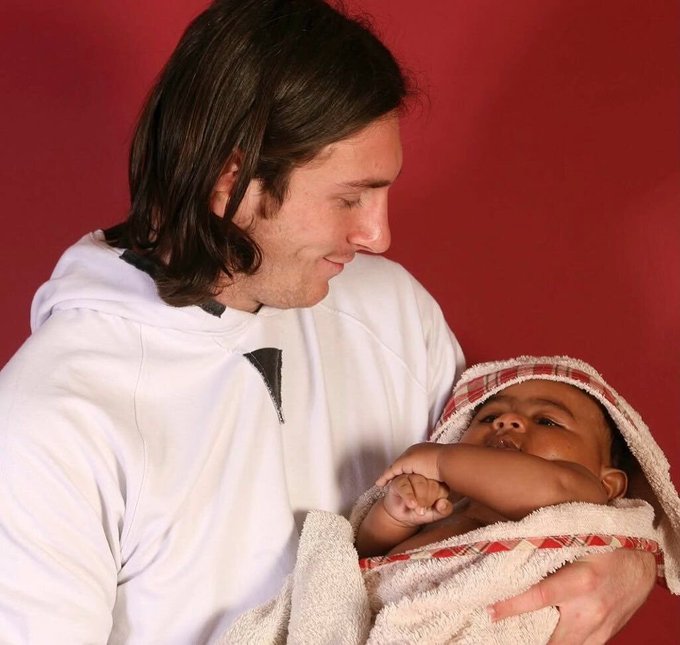 A man with long hair in a white hoodie cradles a baby wrapped in a red and white hooded towel, gazing down tenderly at the child against a deep red background. The juxtaposition of his warm, loving expression with the post's caption humorously frames the image as if his affectionate stare mirrors how he'd lovingly look at his wallet before financial destruction strikes.