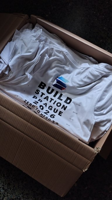 An open cardboard box containing white apparel with 'BUILD STATION PRAGUE 2026 ZERO TO HERO' printed on the fabric.