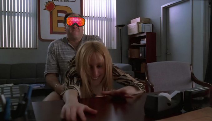 Movie still of a man with glowing red heart-shaped sunglasses standing behind a blonde woman leaning over a desk in an office with a large 'E' logo on the wall.