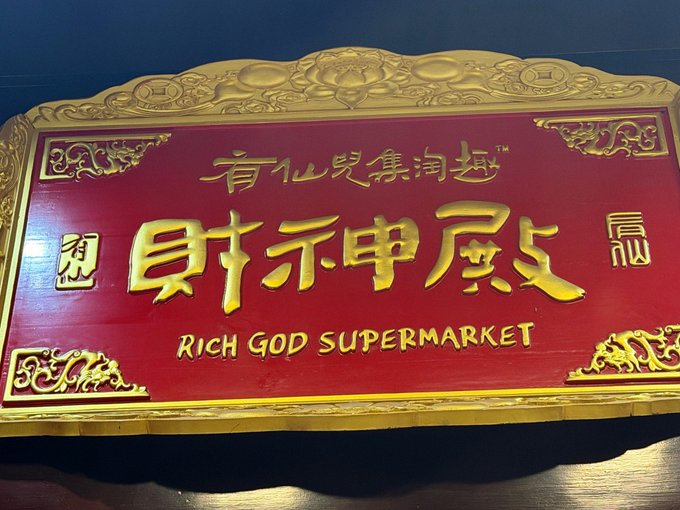 An ornate red and gold plaque displays the Chinese characters "財神殿" alongside the English text "RICH GOD SUPERMARKET" and smaller Chinese text "有仙兒集淘趣," matching the "RICH GOD" reference with the establishment's full name.