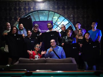 A group of people pose behind a pool table, holding up dark merchandise t-shirts at what appears to be a promotional event.